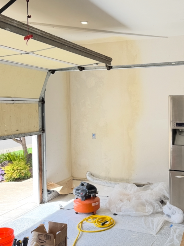 A garage scene showing a refrigerator and a hose, with shelves and tools visible in the background, indicating a workspace